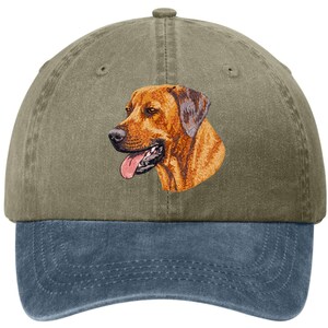 May include: A two-tone baseball cap featuring an embroidered image of a dog's head. The top of the cap is olive green, while the brim is a faded blue. The dog is brown with a pink tongue.