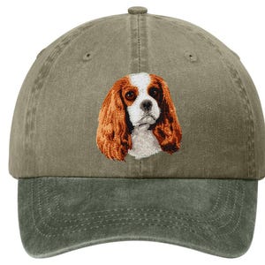 May include: Olive green baseball cap with a two-tone design. The cap features an embroidered Cavalier King Charles Spaniel dog portrait in shades of brown, white, and black. The brim is a darker shade of green.