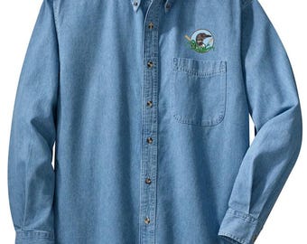 May include: Light blue denim button-down shirt with long sleeves. Features a button-down collar, a chest pocket, and a small embroidered patch on the left chest. The patch depicts a bird in a circular design. The shirt has a classic, casual style.