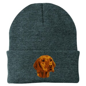 May include: A dark gray knit beanie featuring an embroidered image of a golden-brown dog's head. The dog's fur is detailed with shading, and the beanie has a folded cuff. This winter hat is a cozy accessory for dog lovers.