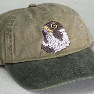 May include: Olive green baseball cap with a dark green brim. The cap features an embroidered image of a falcon's head in shades of brown, white, and yellow. The cap is made of a soft, textured fabric.