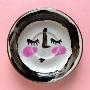 Könnte beinhalten: Eine weiße Keramik-Untertasse mit einem schwarz bemalten Rand und einem handbemalten Gesicht mit geschlossenen Augen, einer Nase und einem Mund. Das Gesicht hat rosa Wangen.