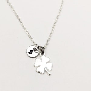 May include: A silver necklace with a four-leaf clover charm and a small round charm with the letter "S" on it.