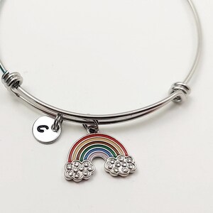 May include: A silver expandable bangle bracelet with a rainbow charm and a letter 'C' charm. The rainbow charm has a red, orange, yellow, green, blue, indigo, and violet design with white clouds.
