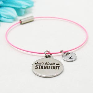 May include: Pink string bracelet with a silver charm that says "don't blend in. STAND OUT." and a silver charm with the letter "K" on it.