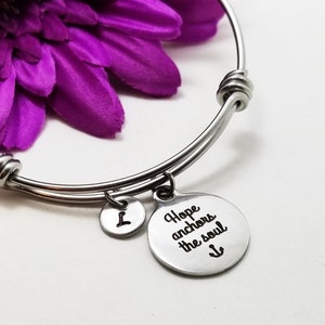 Könnte beinhalten: Ein silbernes Armreif-Armband mit einem Anhänger, auf dem "Hope anchors the soul" und ein Anker-Symbol stehen. Das Armband liegt auf einem lila Blumenhintergrund.