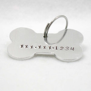 May include: Silver bone-shaped dog tag with a ring for attaching to a collar. The tag has the text "XXX-XXX-1234" engraved on it.