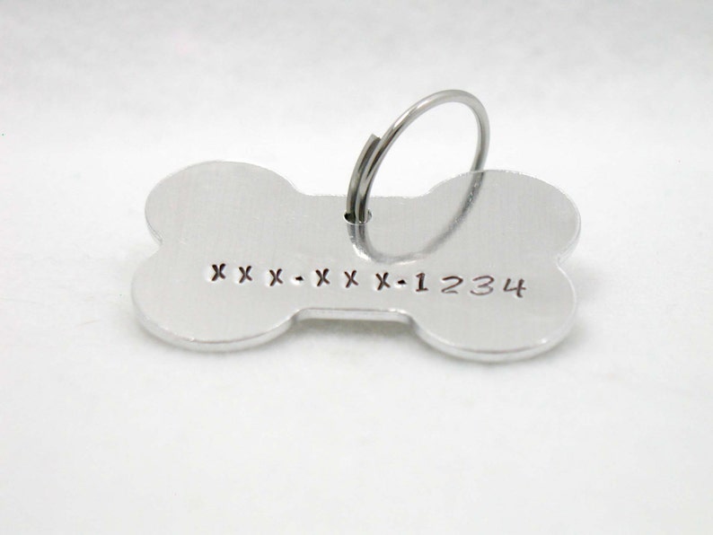 May include: Silver bone-shaped dog tag with a ring for attaching to a collar. The tag has the text "XXX-XXX-1234" engraved on it.