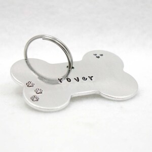 May include: Silver bone-shaped dog tag with the name "Rover" engraved on it. The tag has three paw prints and a ring for attaching to a collar.