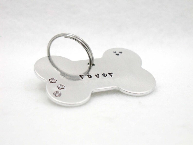 May include: Silver bone-shaped dog tag with the name "Rover" engraved on it. The tag has three paw prints and a ring for attaching to a collar.