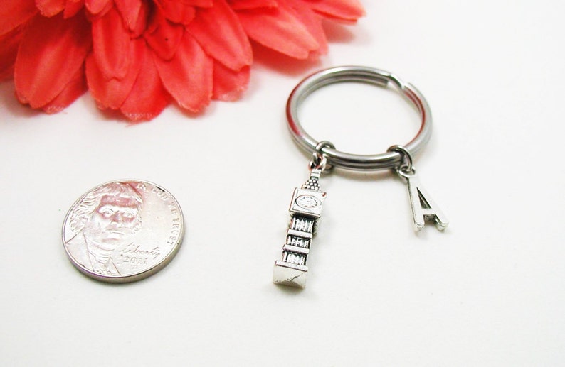 May include: A silver-colored keychain with a Big Ben charm and the letter "A" charm. The keychain is on a white surface with a red flower and a coin in the background.