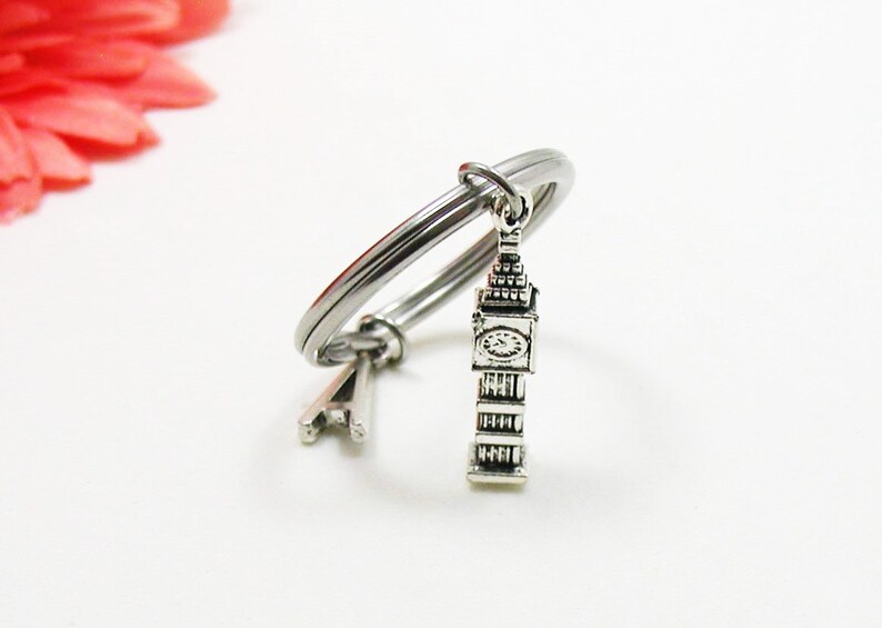 May include: A silver-colored bracelet with a Big Ben charm. The charm is a detailed miniature of the iconic clock tower. The bracelet is on a white surface, with a blurred pink flower in the background.