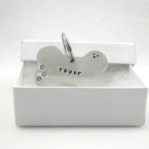 May include: Silver bone-shaped dog tag with the name "rover" engraved on it. The tag has three paw prints and two hearts engraved on it. The tag is in a white box.