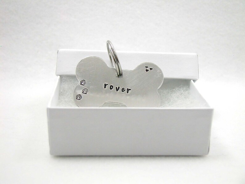 May include: Silver bone-shaped dog tag with the name "rover" engraved on it. The tag has three paw prints and two hearts engraved on it. The tag is in a white box.