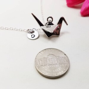 May include: Silver origami crane necklace with a silver initial charm. The initial charm is a letter D. The necklace is shown with a US penny for size comparison.