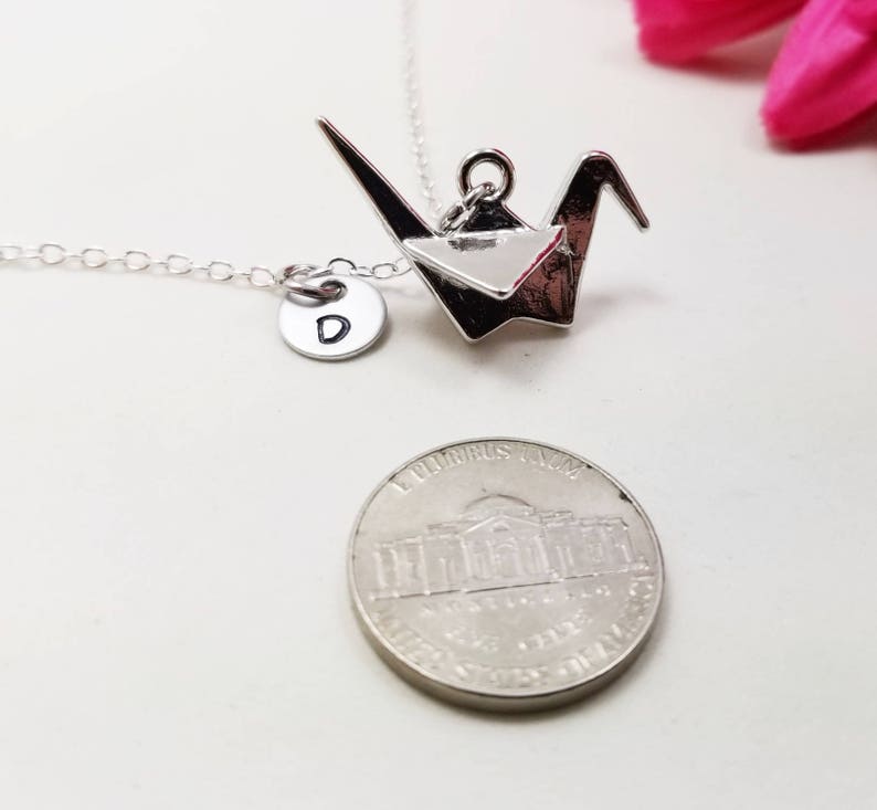 May include: Silver origami crane necklace with a silver initial charm. The initial charm is a letter D. The necklace is shown with a US penny for size comparison.