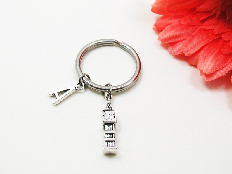 May include: Silver-toned keychain featuring a Big Ben charm and an initial "A" pendant. The keychain has a round ring and is likely made of metal. A decorative accent of a red-orange flower is in the background.