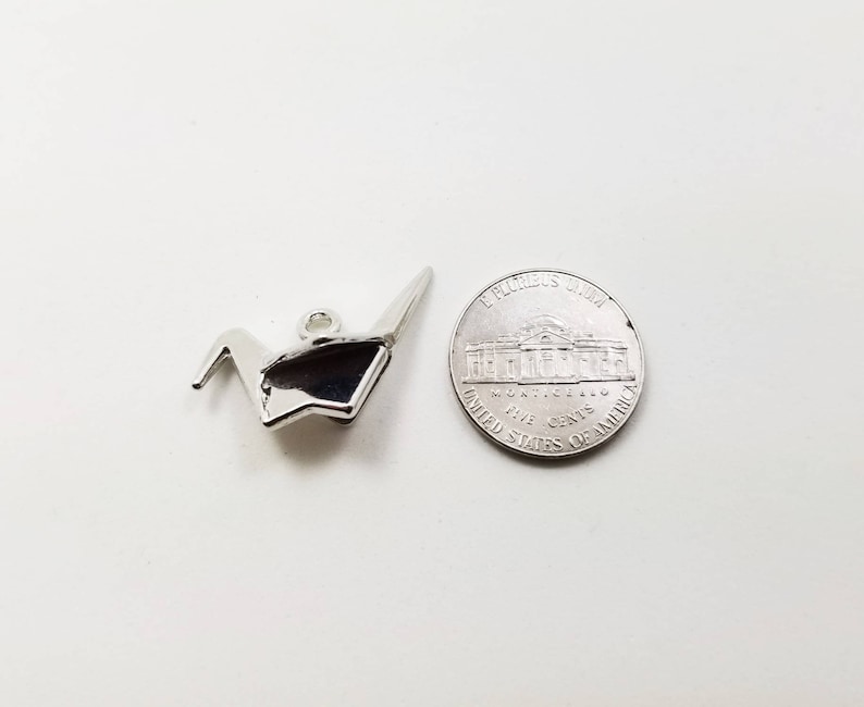 May include: A silver origami crane charm, next to a US five-cent coin.