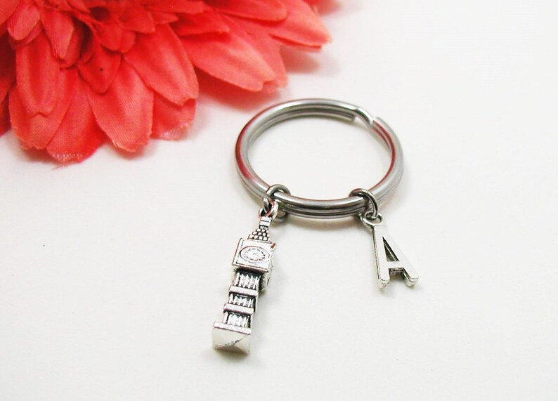 May include: Silver-toned keychain featuring a Big Ben charm and the letter "A" pendant. The keychain has a large, circular ring. A red flower is in the background.