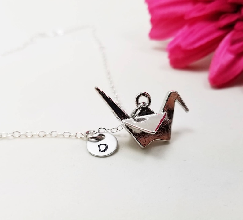 May include: Silver necklace with a silver origami crane charm and a silver disc charm with the letter 'D' engraved on it.