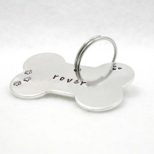 May include: Silver bone-shaped dog tag with the name "Rover" engraved on it. The tag has three paw prints engraved on the left side and a ring for attaching to a collar.