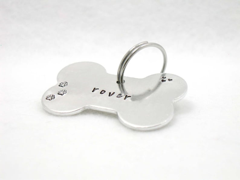 May include: Silver bone-shaped dog tag with the name "Rover" engraved on it. The tag has three paw prints engraved on the left side and a ring for attaching to a collar.