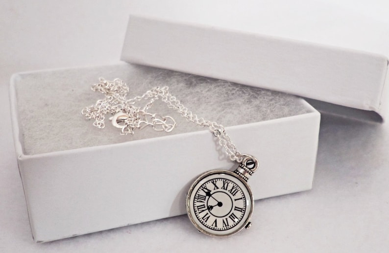 May include: A silver necklace with a round clock pendant. The clock face has Roman numerals and black hands. The necklace is in a white gift box.