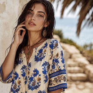 Könnte beinhalten: Ein cremefarbenes Tunika-Oberteil mit blauem Blumenmuster und V-Ausschnitt. Die Ärmel und der Ausschnitt sind mit einem blau-braunen Rand versehen. Das Model trägt goldfarbene Ohrringe und Halsketten.