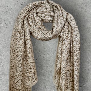 May include: A cream-colored scarf with a repeating pattern of gold-colored leaf-like shapes. The scarf is draped, showing its texture and design. The background is a plain, light gray wall.