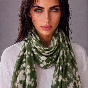 May include: A lightweight scarf featuring a mottled pattern of deep olive green and off-white patches. The fabric is draped around the neck, showcasing a soft, semi-sheer texture with an organic, cloud-like design across the entire surface.