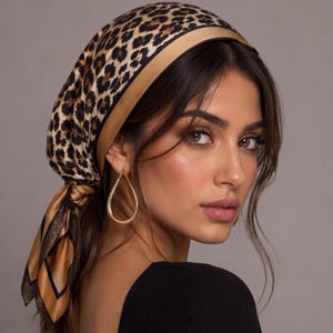 Puede incluir: Un pañuelo cuadrado con estampado de leopardo en marrón y negro y un borde liso de color tostado. El pañuelo está atado en la nuca y muestra un acabado suave y satinado. Un pendiente dorado en forma de lágrima cuelga del lóbulo de la oreja.