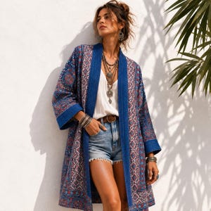 May include: A long-sleeved open-front kimono jacket featuring a blue, red, and white geometric pattern, worn over a plain white camisole and light blue denim shorts with a brown leather belt, accessorised with layered silver necklaces and bracelets.