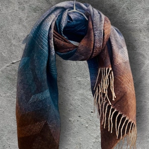 May include: A large scarf with a plaid pattern in shades of blue, brown, and gray, hanging on a white hanger. The scarf has a fringed edge and is made of a soft, woven material. The scarf is a winter accessory.