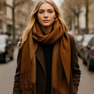 Op de afbeelding: Een vrouw draagt een bruine sjaal en jas. De sjaal heeft een warme bruine kleur met een gefranjerde rand. De jas is een donkerdere bruintint. De vrouw heeft lang blond haar en kijkt recht in de camera.
