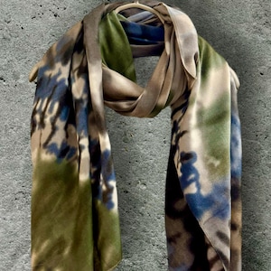 May include: A draped scarf with a watercolor-style pattern in shades of blue, green, and brown. The scarf is made of a soft, flowing fabric and is displayed on a hanger.