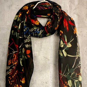 Può includere: Una sciarpa nera con un colorato motivo floreale. La sciarpa presenta una varietà di fiori e foglie nei toni dell'arancione, giallo, blu e verde. È appesa a una gruccia bianca su uno sfondo grigio.