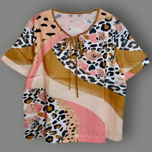 May include: A short-sleeved top with a V-neck and tie closure. The top features a colourful pattern with leopard print, floral designs, and abstract shapes in shades of brown, pink, and white. The neckline and tie are brown.