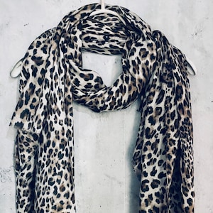 May include: A leopard print scarf, draped over a white hanger. The scarf features a repeating pattern of brown spots on a cream background. The fabric appears lightweight and flowing, suitable for various styles.