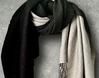 Può includere: Una sciarpa reversibile con un lato nero a tinta unita e un lato beige chiaro. Il tessuto ha un aspetto intrecciato e testurizzato con un sottile luccichio. Un'estremità della sciarpa presenta un dettaglio a frange con fili neri e beige.