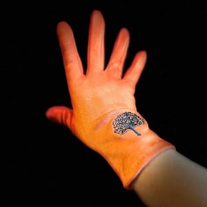 May include: Bright orange glove featuring a black tree design on the palm. The glove is made of a soft material and is set against a black background. The tree design is detailed and stylized.