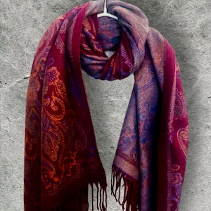 Op de afbeelding: Bordeaux sjaal met een paisley patroon in tinten oranje, paars en blauw. De sjaal heeft een franjerand en is over een witte hanger gedrapeerd. De sjaal is gemaakt van een zacht, soepel materiaal.