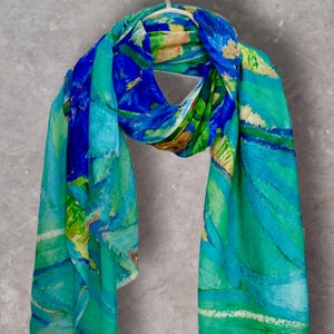Puede incluir: Un pañuelo ligero con un patrón abstracto de pinceladas en azul vibrante, turquesa y verde lima. La tela muestra una estética pictórica con degradados de color mezclados y bordes suaves y deshilachados, colgado sobre una percha blanca.