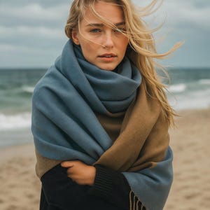 May include: A woman wearing a large, two-tone scarf on a beach. The scarf is blue and tan, with a fringed edge. The woman is wearing a black sweater. The background shows the sea and a cloudy sky.