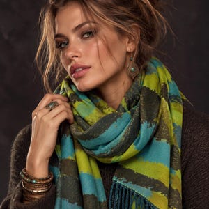Op de afbeelding: Een sjaal met horizontale strepen in limoengroen, teal en donkerbruin met subtiele metallic gouden accenten. De sjaal heeft een gefranjerde rand en wordt gedragen met een donkerbruine trui, een ring met een groene steen en gestapelde armbanden.