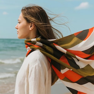 May include: A colorful scarf with geometric patterns in shades of orange, green, black, and white is draped around the neck. The scarf is blowing in the wind against a backdrop of a beach and blue sky. The person is wearing a white shirt.
