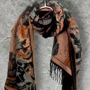 Op de afbeelding: Een sjaal met een bloemenpatroon in zwart, oranje en bruin. De sjaal is over een hanger gedrapeerd, met de franjes en het ingewikkelde patroon. De kleuren en het ontwerp suggereren een herfst- of winteraccessoire.