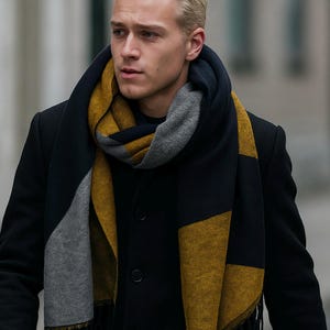 Peut inclure: Écharpe à motifs géométriques dans les tons bleu marine, jaune moutarde et gris. Elle est portée autour du cou d'une personne vêtue d'un manteau noir. Les extrémités sont effilochées.