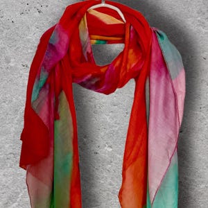 Puede incluir: Una bufanda vibrante y colorida con efecto acuarela. La bufanda presenta un degradado de tonos rojos, naranjas, rosas, verdes y azules. Está colgada de una percha blanca sobre un fondo gris. La bufanda está hecha de una tela ligera y fluida.