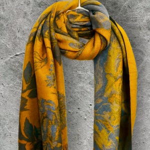 May include: Mustard yellow scarf with a floral pattern in shades of gray and blue. The scarf is draped over a white hanger against a gray background. The scarf's design features botanical elements.