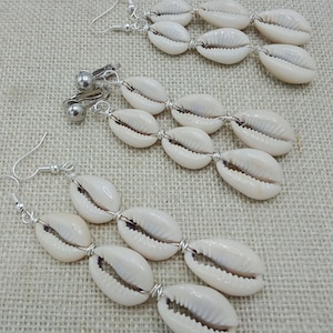 May include: Three pairs of seashell earrings. Each earring features three white, oval-shaped seashells strung together with silver-colored wire. Two pairs have hook closures, and one pair has clip-on closures. The earrings rest on a textured, beige surface.